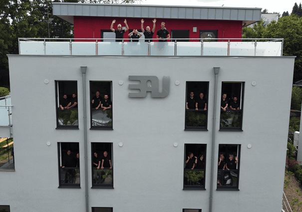 Belegschaft von BAU-Arbeitsschutz steht auf dem Balkon des Arbeitsgebäudes und winkt freundlich in die Kamera