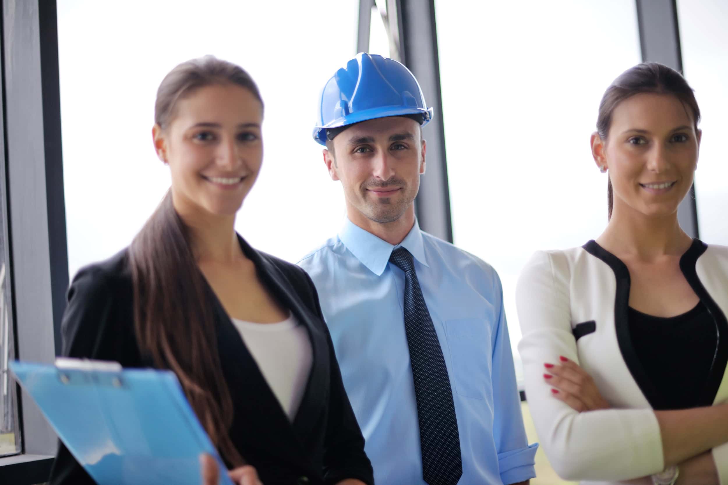 Drei Business Leute stehen in einem modernen Büro, die mittlere Person trägt einen Arbeitshelm